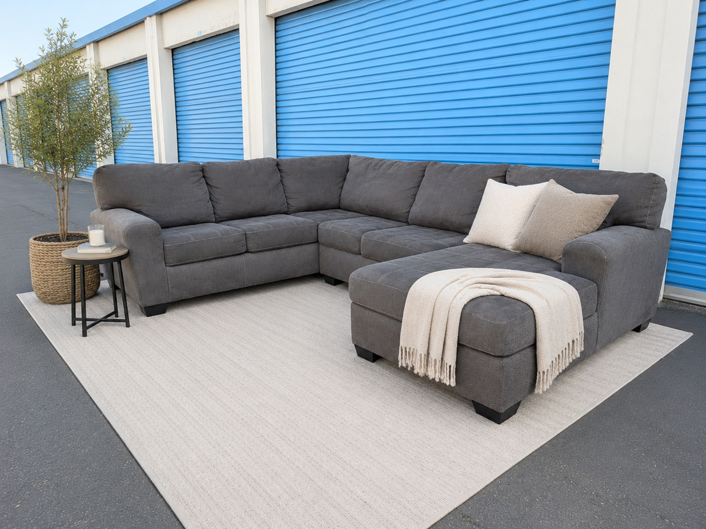 A dark gray sectional sofa with a chaise lounge and two cream throw pillows, styled with a cream fringed throw blanket on a light beige rug. The sofa is set outdoors in front of a building with blue roll-up garage doors, next to a small side table with a potted tree.